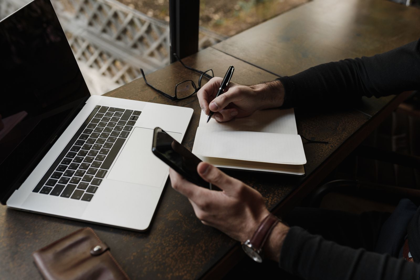 Hands writing in a notebook beside a smartphone and laptop